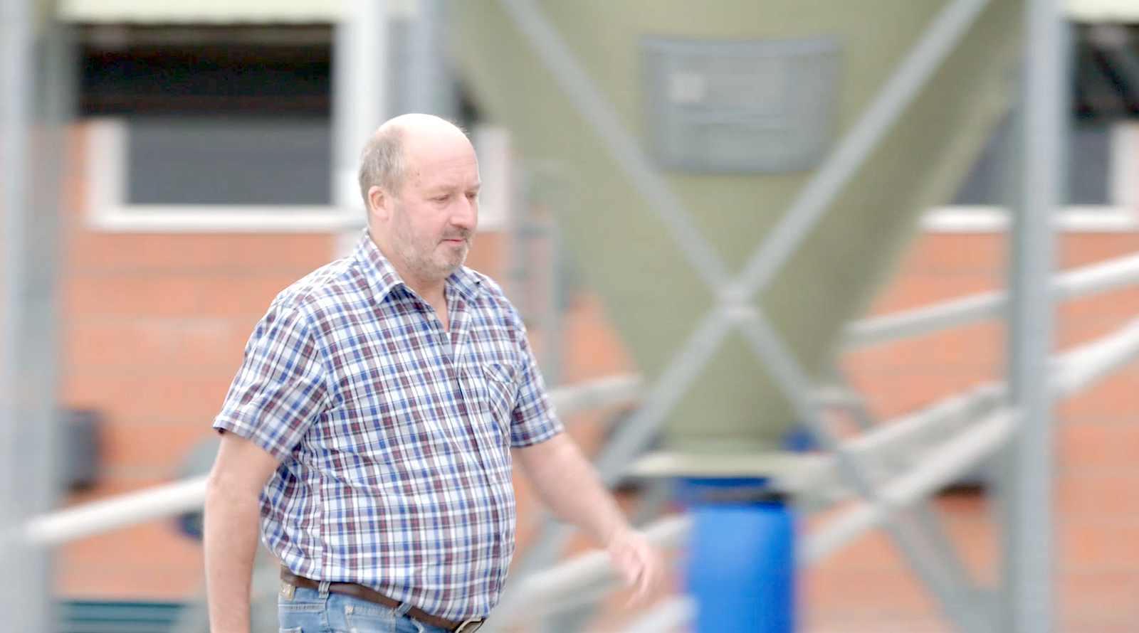 Hähnchenhalter Diedrich Dammann läuft im karierten Hemd und Jeans an seinem Stall vorbei. Der Hintergrund ist unscharf. Es ist ein Futtersilo erkennbar. 