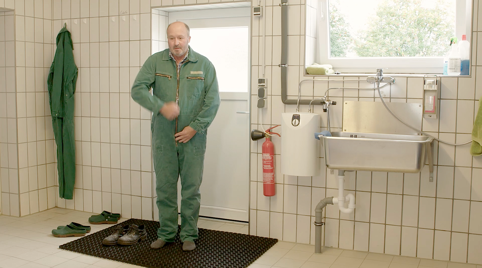 Der Hähnchenhalter Diedrich Dammann macht den Reißverschluss seines grünen Schutzanzuges zu. Er steht auf Socken auf einer Fußmatte im Vorraum seines Hähnchenstalls. 