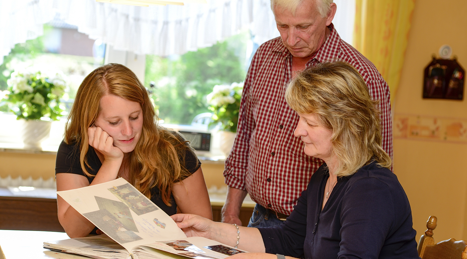 Hähnchenhalterin Ramona Harkers sitzt mit ihrer Mutter am Küchentisch und schaut sich ein Fotoalbum an. Ihr Vater, der ebenfalls Hähnchenhalter ist, steht zwischen den beiden. 